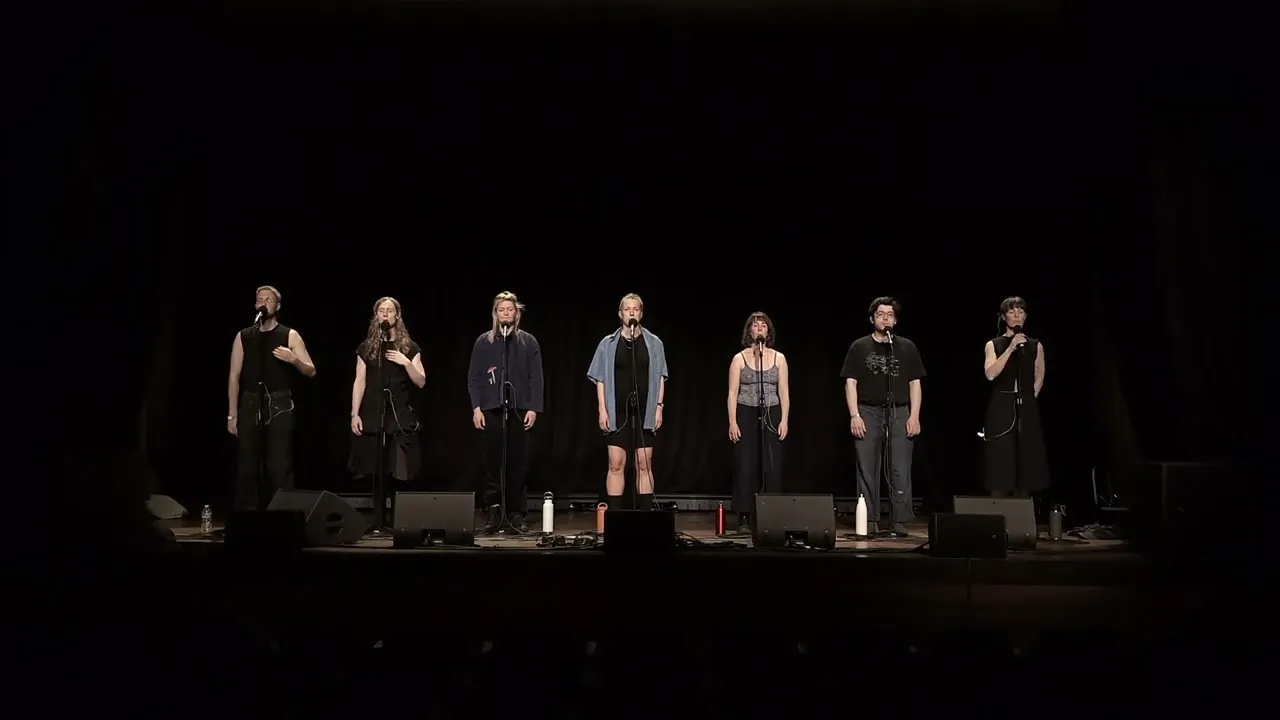 Seven singers standing in a line at microphones on a dark stage at Bristol Beacon