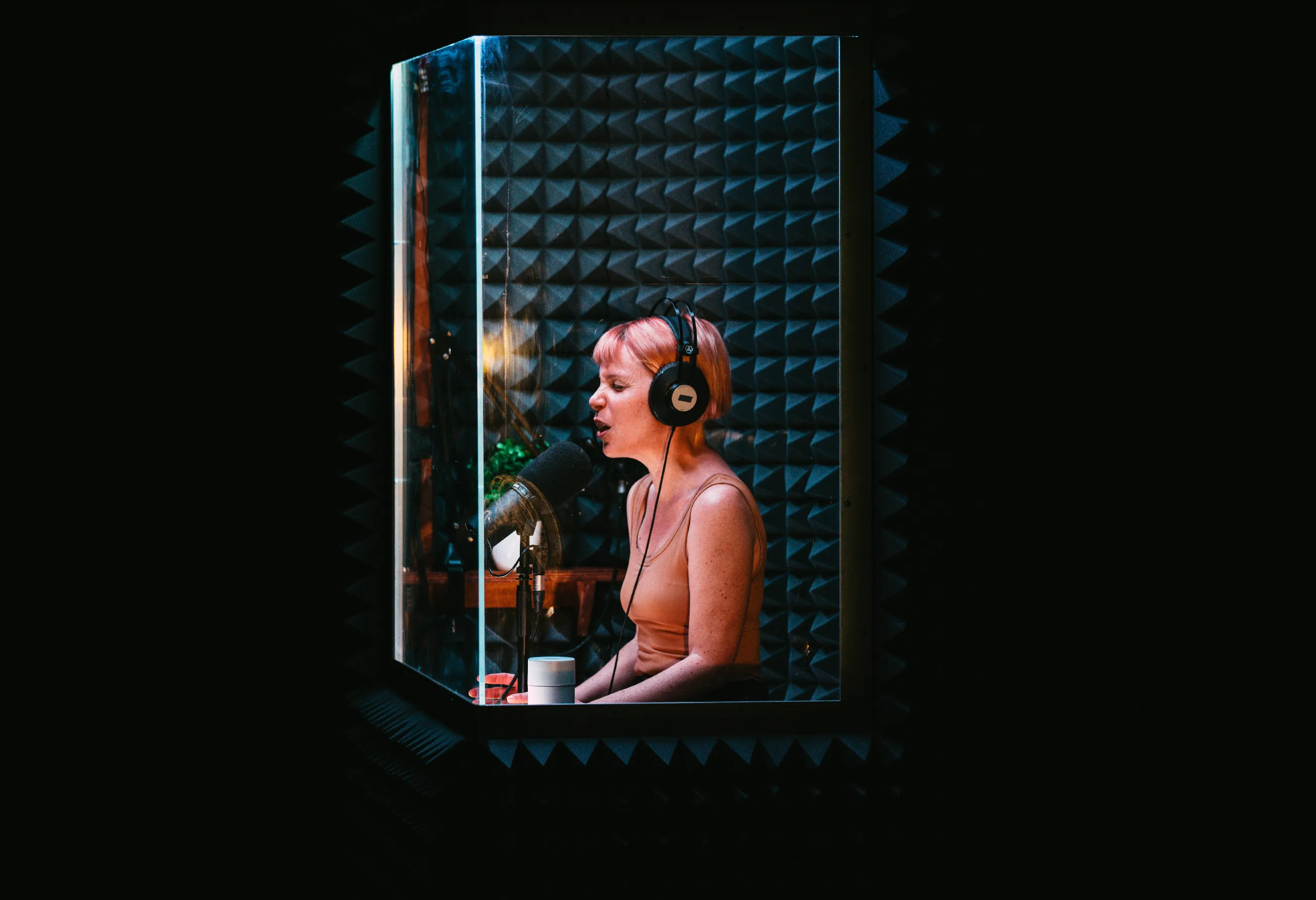 Gemma Paintin in headphones speaking into a microphone inside a glass-fronted vocal booth lined with acoustic foam